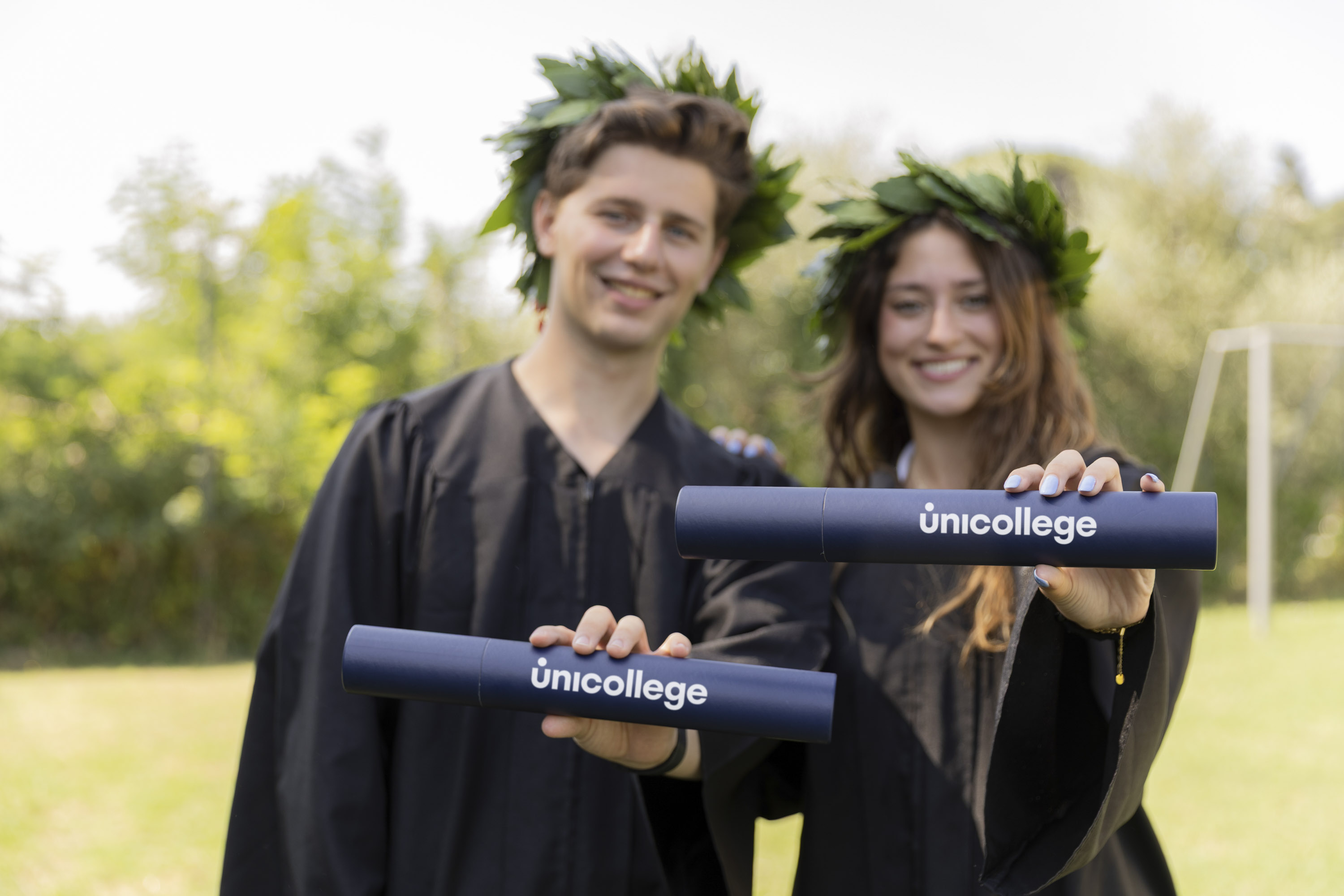 Two Unicollege graduates wearing laurel wreaths and holding their diplomas