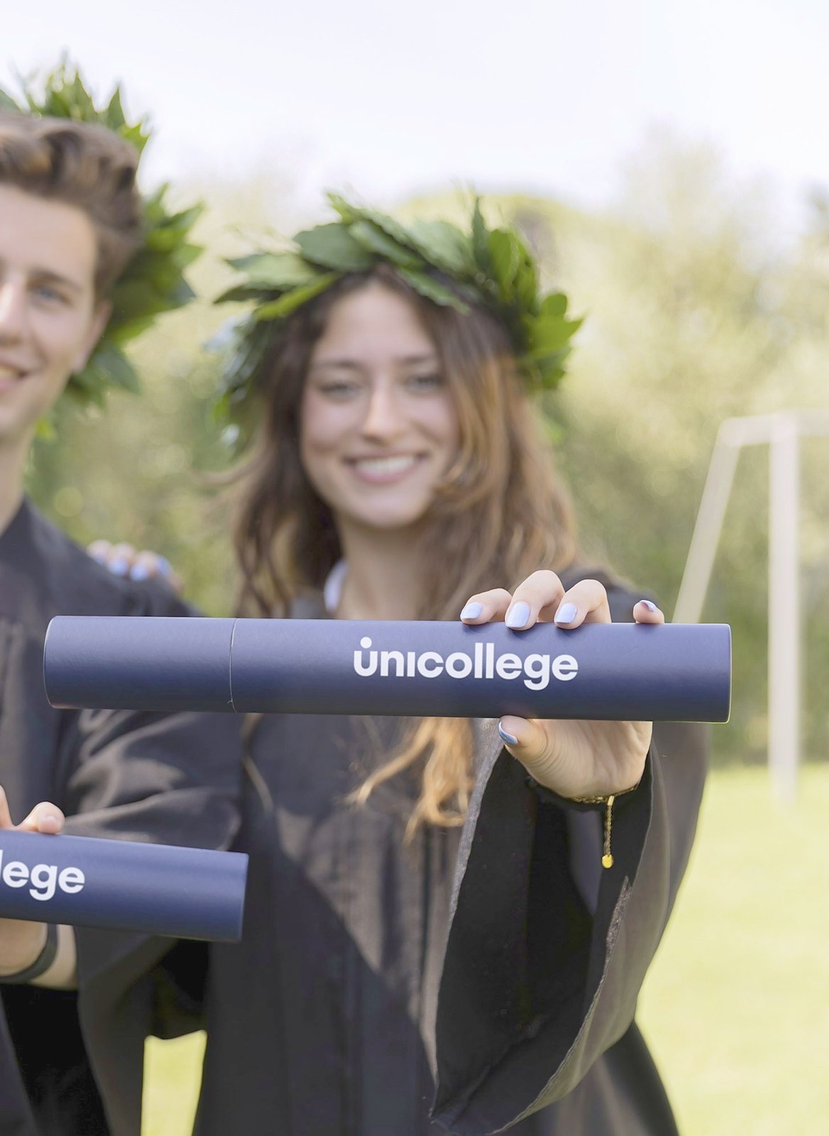 Two graduates in gowns and laurel wreaths holding their Unicollege diploma tubes