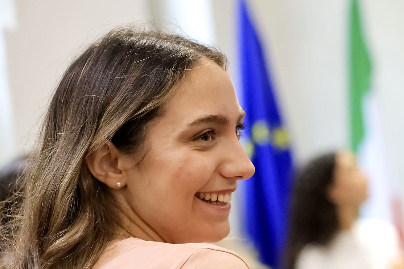 A smiling Unicollege student in class, with the Italian and European flags behind her, Turin campus