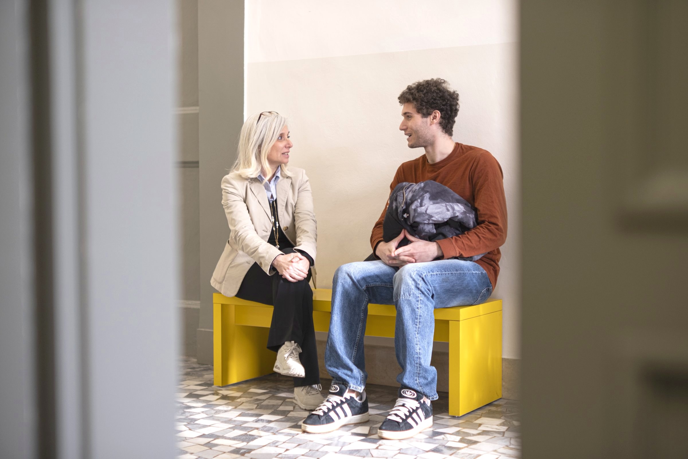 Two students sitting on the yellow benches in a campus corridor